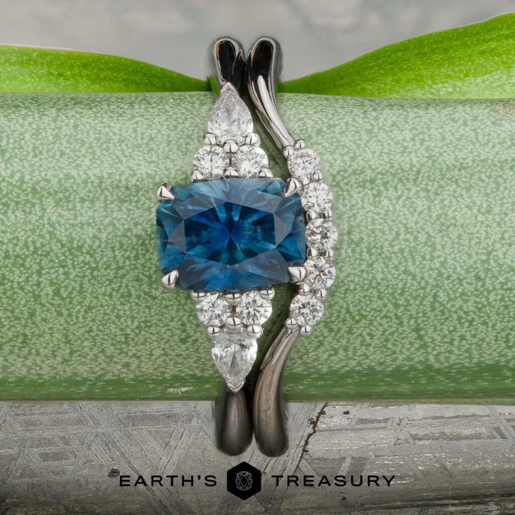 The Patterson, Sean ring features a square-cut blue gemstone with diamond accents on a silver band, displayed upright on a green leaf, evoking the elegance of women's wedding bands.