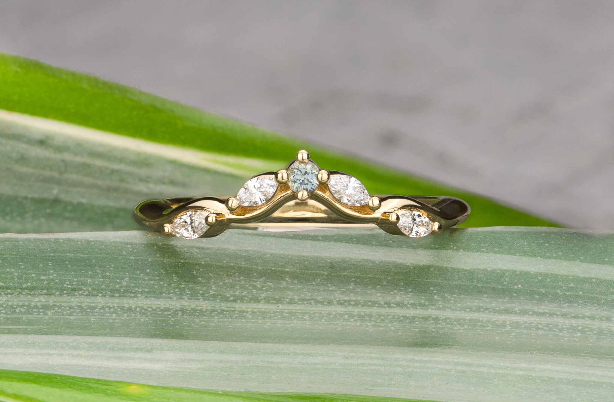 Stacked from 25 images (Method=B, R=11, S=7): a gold ring with five marquise and round diamonds in a curved design, shown on green striped leaves and a gray background—a unique custom wedding band for elegant style.