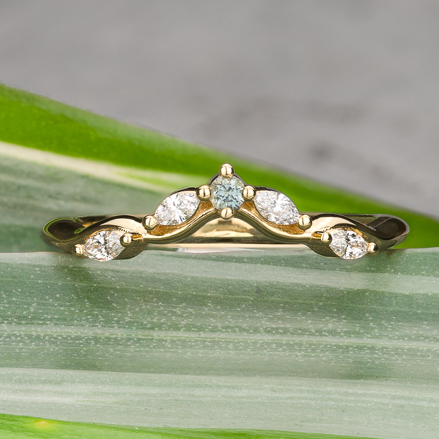 The Womens-Band-sqr is a gold ring with a curved band, set with five marquise and round white gemstones—an elegant, custom wedding band displayed on green leaves.