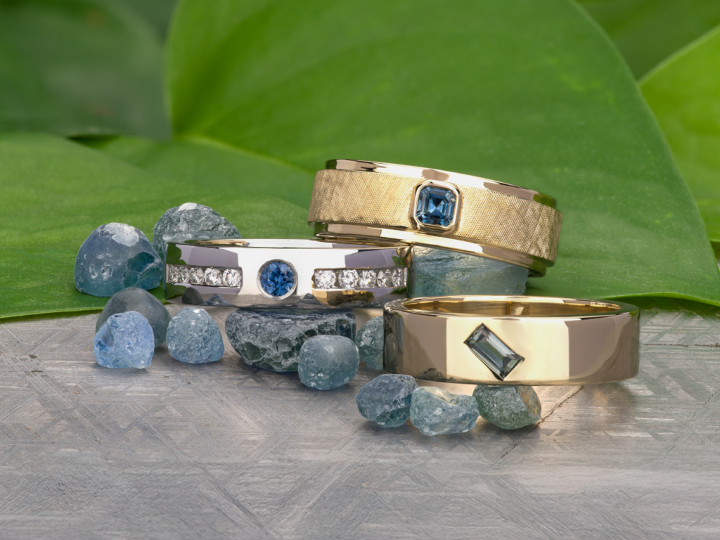The mens-unisex-scaled-1-1024x768 wedding bands in gold and silver, set with blue gemstones, are shown among scattered blue stones on a textured surface with green leaves in the background.