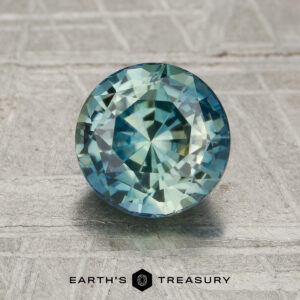 A round, faceted 2.03-carat unheated blue-green sapphire stands upright on a textured gray surface with "EARTH'S TREASURY" below.