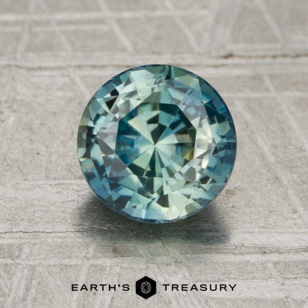 2.03-Carat Spruce Green Madagascar Sapphire (Unheated) A round, faceted 2.03-carat unheated blue-green sapphire stands upright on a textured gray surface with "EARTH'S TREASURY" below.