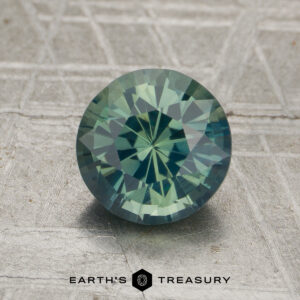 A round, faceted 1.78-carat unheated sapphire with green and blue hues rests on a textured surface. The "Earth's Treasury" logo appears at the bottom.