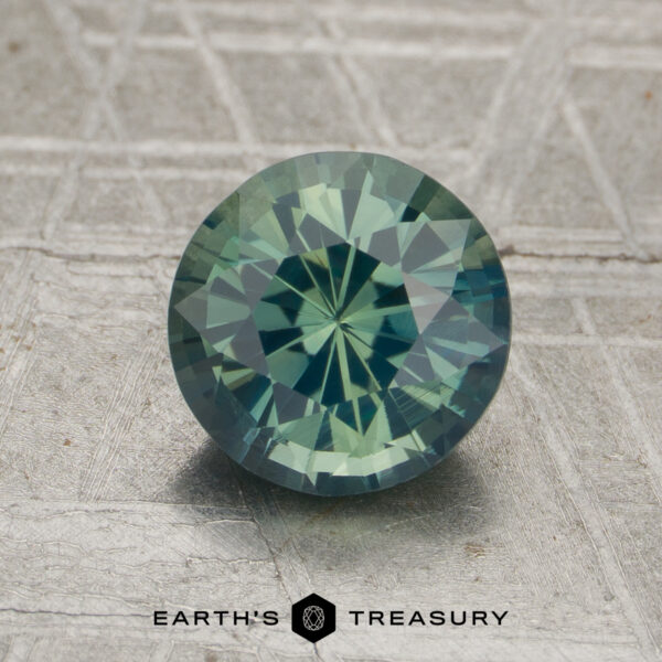 A round, faceted 1.78-carat unheated sapphire with green and blue hues rests on a textured surface. The "Earth's Treasury" logo appears at the bottom.