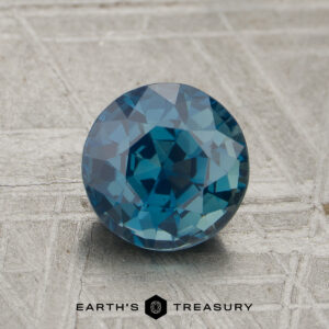 A round, faceted 1.76-carat unheated sapphire is displayed on a textured gray surface with "EARTH'S TREASURY" text and logo at the bottom.