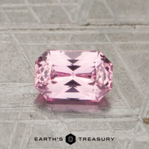A 1.15-carat unheated pink sapphire with a rectangular, faceted cut rests on a textured gray metallic surface, with the text "EARTH'S TREASURY" beneath it.