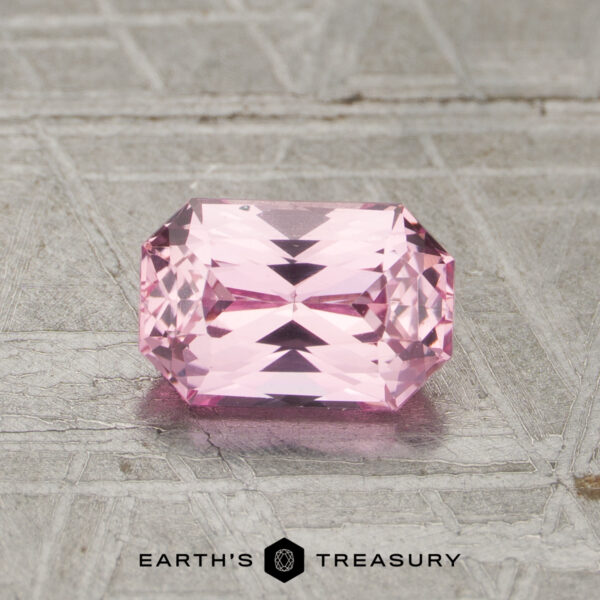 A 1.15-carat unheated pink sapphire with a rectangular, faceted cut rests on a textured gray metallic surface, with the text "EARTH'S TREASURY" beneath it.
