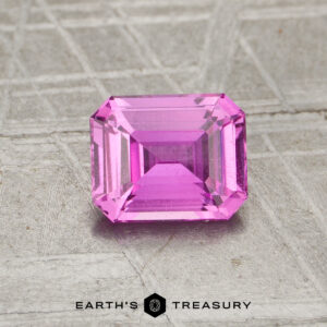A rectangular, step-cut pink 1.15-carat unheated sapphire rests on a textured gray surface with the text "EARTH'S TREASURY" at the bottom.