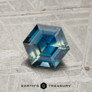 A 1.12-carat sapphire with geometric facets rests on a textured gray surface. The Earth's Treasury logo and text appear at the bottom.