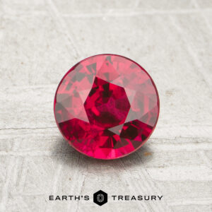 A 1.12-carat round, faceted ruby is displayed on a textured gray surface with the "Earth's Treasury" logo beneath it.