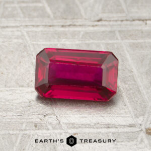 A 1.13-carat, emerald-cut ruby rests on a textured surface with "EARTH'S TREASURY" and a hexagonal logo below.