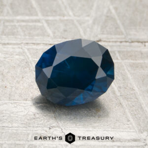 A 1.57-carat heated Montana sapphire, cut in a faceted oval shape, rests on a textured surface with the "EARTH'S TREASURY" name and logo visible at the bottom.