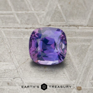 A 1.04-carat unheated sapphire with a cushion cut and blue-violet hue is showcased on a textured metallic surface, with "EARTH'S TREASURY" displayed at the bottom.
