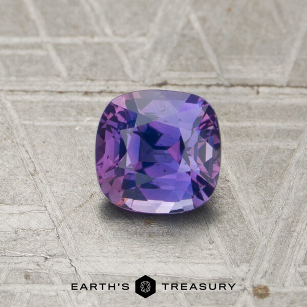 A 1.04-carat unheated sapphire with a cushion cut and blue-violet hue is showcased on a textured metallic surface, with "EARTH'S TREASURY" displayed at the bottom.