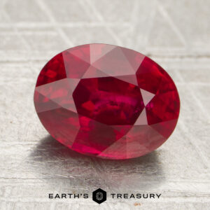 A 2.51-carat faceted oval ruby rests on a textured surface, with "EARTH'S TREASURY" and a hexagonal logo displayed at the bottom.