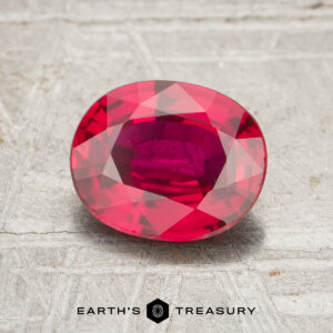 A 1.24-carat oval-cut ruby with a vivid red hue is displayed on a textured light gray surface, featuring "Earth's Treasury" at the bottom.