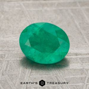A faceted oval green 7.60-carat emerald gemstone displayed on a textured metallic surface, with the "Earth's Treasury" logo and text at the bottom.