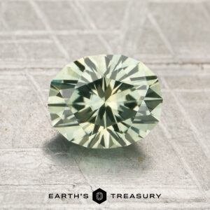 A 1.56-carat heated Montana Sapphire with an oval cut in pale green rests on a textured gray surface, featuring the Earth's Treasury logo at the bottom.
