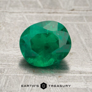 A 1.10-carat faceted green emerald gemstone rests on a textured gray surface, with the "Earth's Treasury" logo and text visible at the bottom.