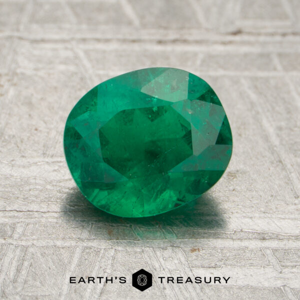 1.10-Carat Rich Green Zambian Emerald (Oiled) A 1.10-carat faceted green emerald gemstone rests on a textured gray surface, with the "Earth's Treasury" logo and text visible at the bottom.