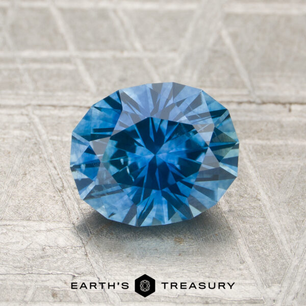 1.54-Carat Medium Blue Montana Sapphire (Heated) A 1.54-carat heated Montana sapphire is displayed on a textured silver-gray surface with "EARTH'S TREASURY" at the bottom.