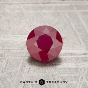A 0.90-carat round, faceted ruby rests on a textured gray surface. The "EARTH'S TREASURY" logo is displayed at the bottom.