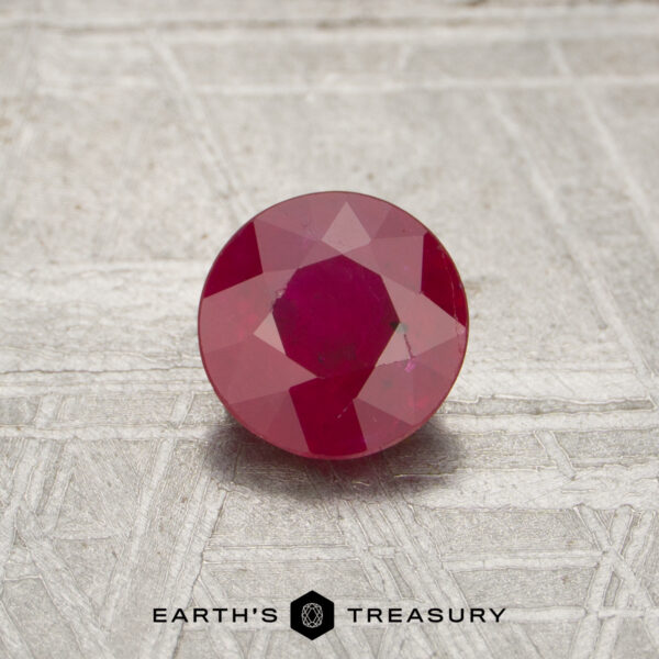 0.90-Carat Mozambique Ruby (Heated) A 0.90-carat round, faceted ruby rests on a textured gray surface. The "EARTH'S TREASURY" logo is displayed at the bottom.