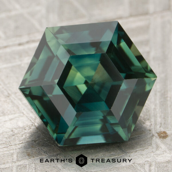 4.53-Carat Spruce Green Madagascar Sapphire A precision-cut hexagonal 4.53-carat sapphire sits on a textured surface, with the Earth's Treasury logo and name displayed at the bottom.