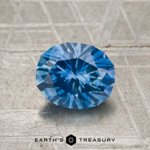 A 1.48-carat heated Montana sapphire is displayed on a textured surface with "Earth's Treasury" and a hexagon logo below.