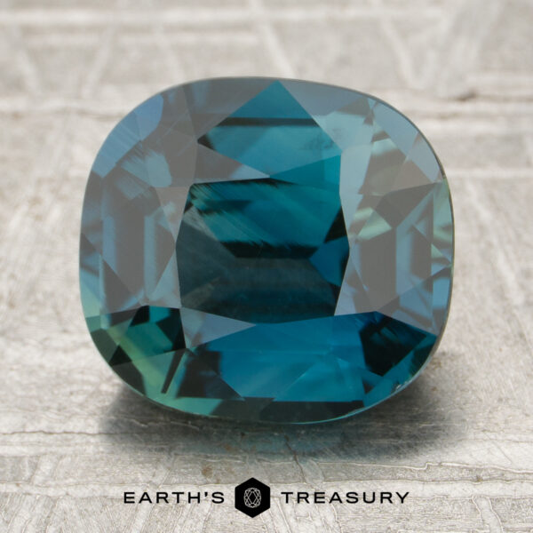 3.51-Carat Teal Madagascar Sapphire A 3.51-carat cushion-cut blue-green sapphire rests on a textured gray surface with "Earth's Treasury" displayed beneath it.
