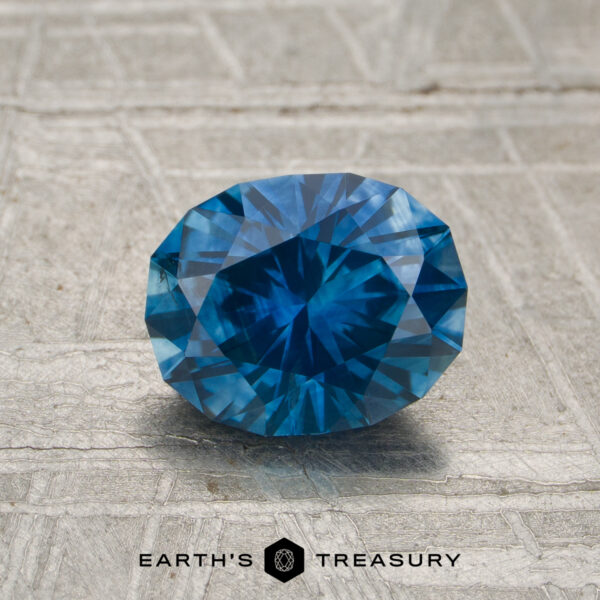 1.44-Carat Rich Teal Montana Sapphire (Heated) A 1.44-carat heated Montana sapphire with blue facets rests on a textured gray surface, with "EARTH'S TREASURY" and a hexagon logo displayed at the bottom.