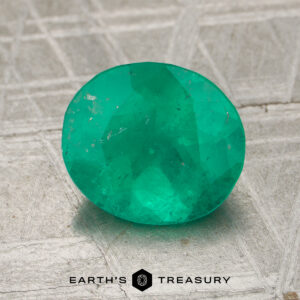 A 2.16-carat oval-cut emerald rests on a textured metallic surface with the words "EARTH'S TREASURY" at the bottom.