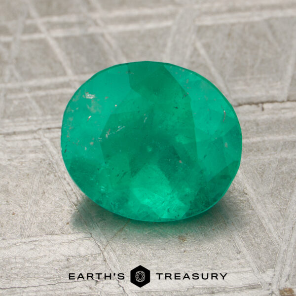 2.16-Carat Rich Green Colombian Emerald (Oiled) A 2.16-carat oval-cut emerald rests on a textured metallic surface with the words "EARTH'S TREASURY" at the bottom.