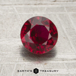 A 1.50-carat round, faceted deep red ruby is displayed on a textured surface with the words "EARTH'S TREASURY" and a hexagon logo beneath it.