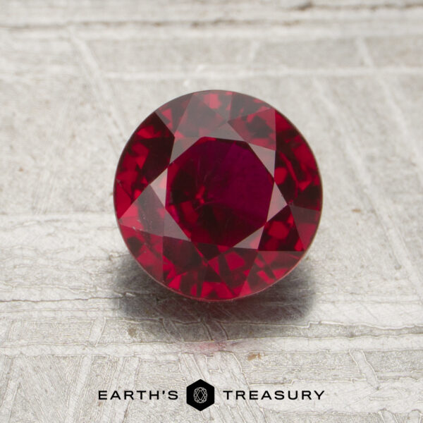 1.50-Carat Mozambique Ruby (Heated) A 1.50-carat round, faceted deep red ruby is displayed on a textured surface with the words "EARTH'S TREASURY" and a hexagon logo beneath it.
