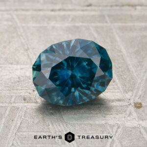 A 1.43-carat heated Montana sapphire, faceted and oval-shaped, rests on a textured surface; "EARTH'S TREASURY" appears below with a geometric logo.