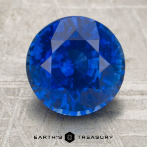 A 3.38-carat round-cut blue sapphire rests on a textured surface, accompanied by the text "EARTH'S TREASURY" and a geometric logo below.