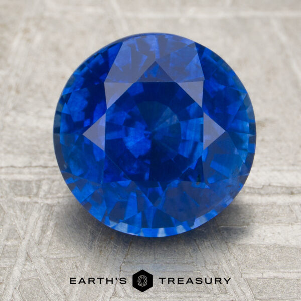 3.38-Carat Sapphire A 3.38-carat round-cut blue sapphire rests on a textured surface, accompanied by the text "EARTH'S TREASURY" and a geometric logo below.