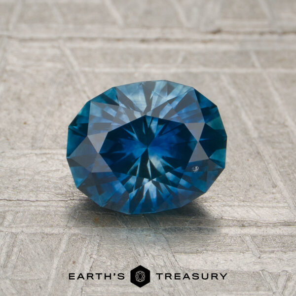 1.42-Carat Rich Blue-Aqua Particolored Montana Sapphire (Heated) A 1.42-carat heated Montana sapphire, oval-cut and blue, displayed on a textured metallic surface with the "EARTH'S TREASURY" logo at the bottom.