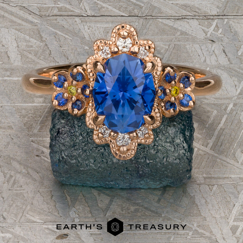 A rose gold ring from Tom, Irene [custom gallery], featuring an oval blue gemstone encircled by small diamonds and blue stones, is displayed on a green mineral with the "Earth's Treasury" logo beneath.