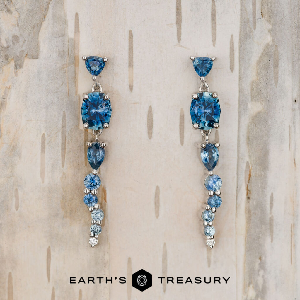 A pair of silver dangle earrings with blue gemstones in assorted shapes and sizes is displayed on light wood, with the Earth’s Treasury logo below. Product: Ball, Justin [custom gallery].