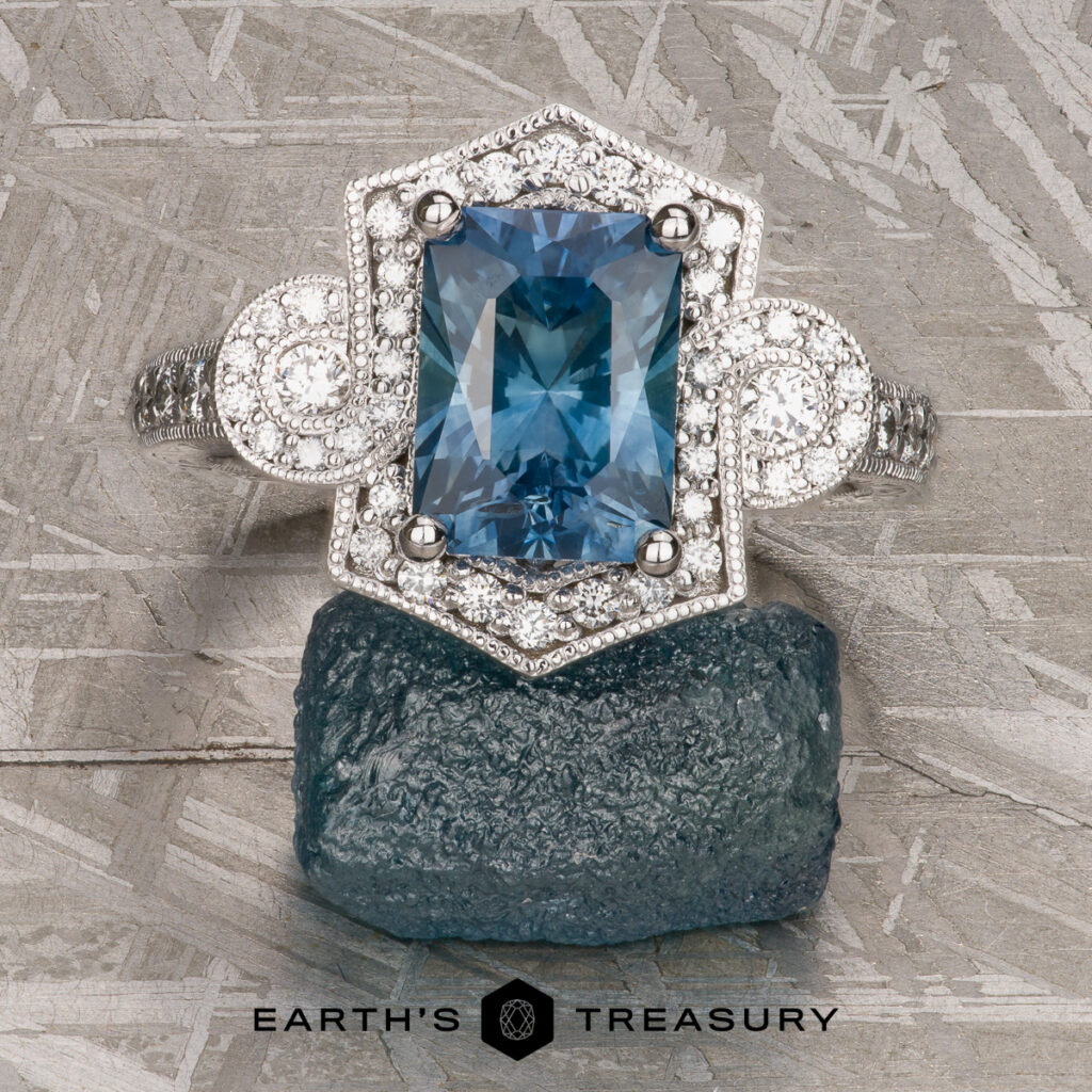 A rectangular blue gemstone ring with diamond accents, from the Gajdocek, Lauren and Tyler custom gallery, is displayed on a dark crystal against a patterned metallic background with the Earth's Treasury logo below.