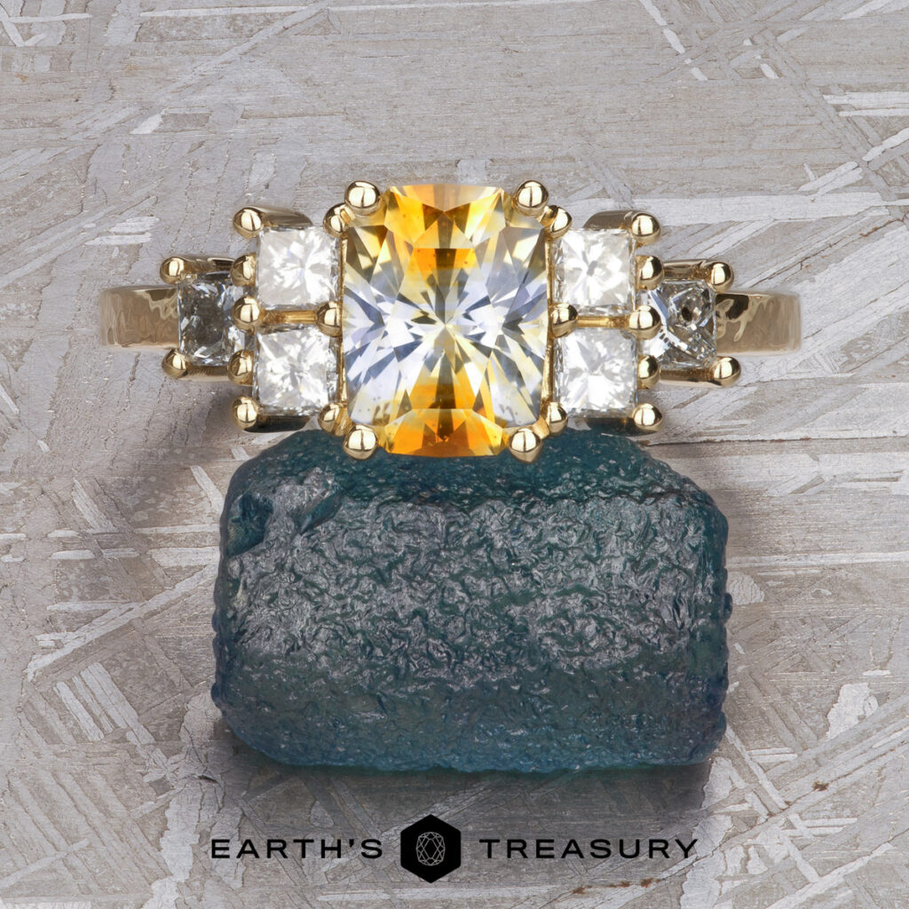A gold ring featuring a rectangular yellow gemstone and clear stones sits atop a blue-green mineral, with the "Jeffries, April and Karas, Joseph [custom gallery]" name displayed below.