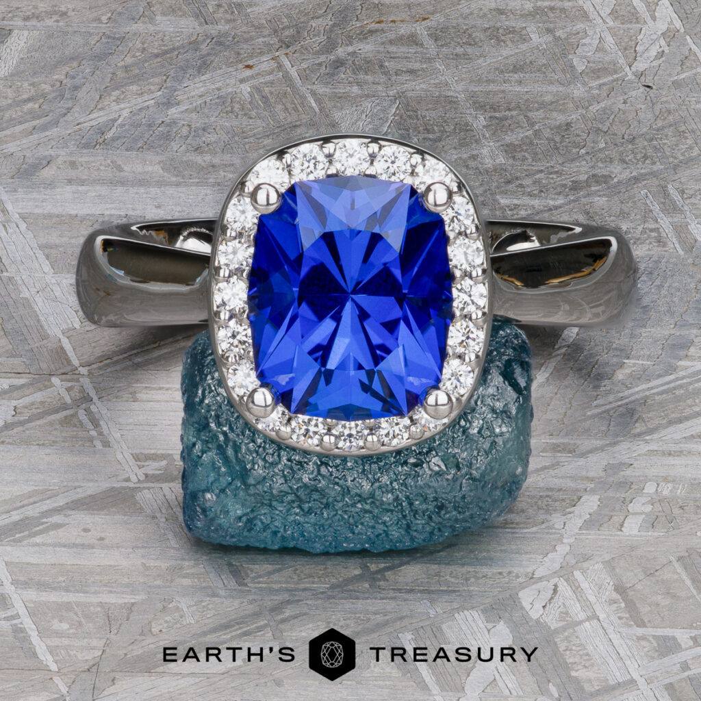 A silver ring with a cushion-cut blue gemstone and small diamonds, from the "Jones, Mike [custom gallery]," is displayed on rough blue stone with the "Earth's Treasury" logo below.