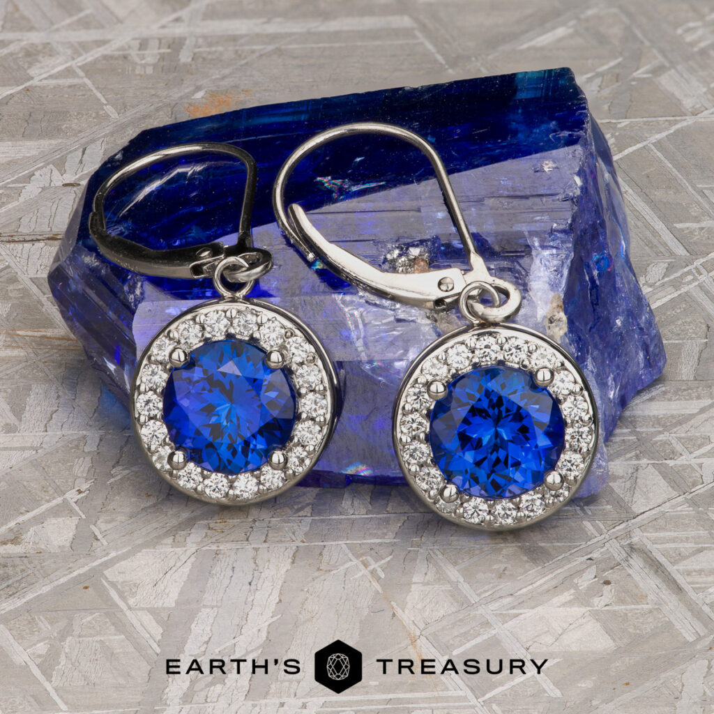 A pair of silver earrings with round blue gemstones and small clear stones set in a circle, displayed on rough blue crystal, from Jones, Mike [custom gallery]. The Earth's Treasury logo appears at the bottom.