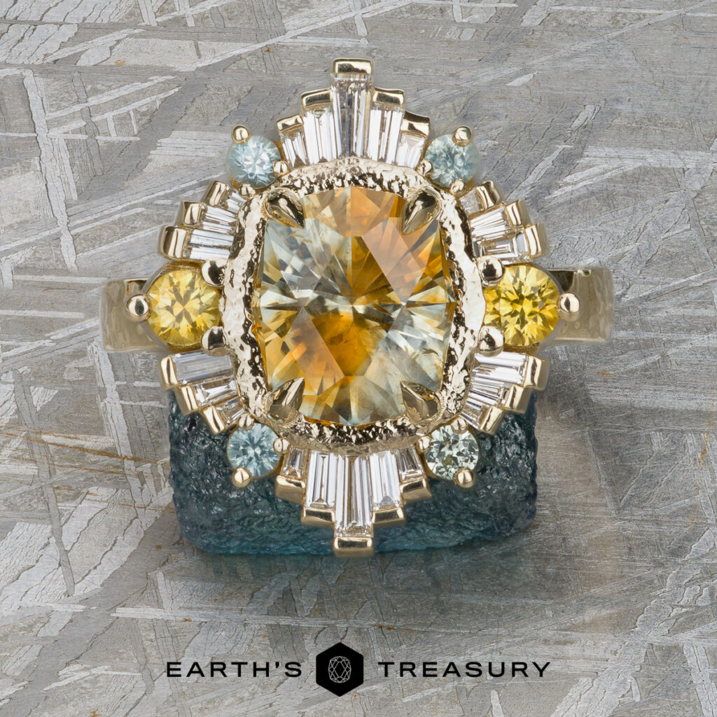 A gold ring, "Dykema, Jennifer [custom gallery]," features a large yellow gemstone center with smaller yellow, blue, and clear stones, displayed on a textured gray surface. "EARTH'S TREASURY" appears below.