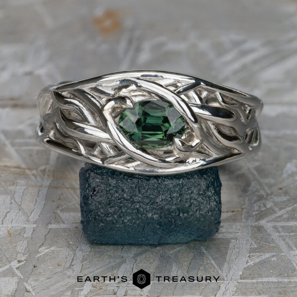 A silver ring with an intricate design and a round green gemstone is displayed on a rough, dark stone. The product, "Zulevic, Athena and Martin, Thomas 3 [custom gallery]," features "EARTH'S TREASURY" text at the bottom.
