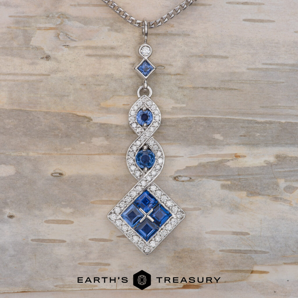 The Clagett, Chance [custom gallery] is a silver pendant necklace featuring blue and white gemstones in geometric and swirling patterns, shown on a light wood background.