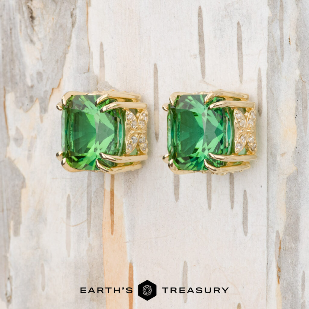 The Krishna Bondili, Meg earrings feature large square green gemstones and small clear stones, shown on a light wooden surface.