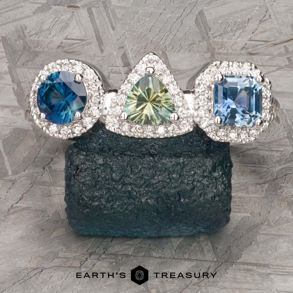 Three gemstone rings from Danielson, Christin [custom gallery] with diamond halos—two blue and one green, each uniquely shaped—are displayed on dark textured stone above the “Earth's Treasury” logo.